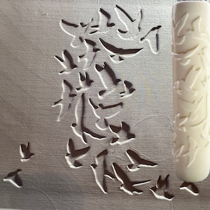 May include: A white rolling pin with a repeating pattern of birds. The rolling pin is used to create an embossed design on a surface. The surface is light grey and has a pattern of birds created by the rolling pin.