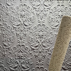 May include: A close-up of a beige rolling pin with a detailed floral pattern. The rolling pin is lying on a textured surface with a similar floral pattern.