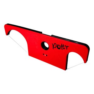 Op de afbeelding: Een rood aardewerk gereedschap met een zwarte rand en een draadsnij-element. De tool heeft een cirkelvormig gat en het woord "Pott'r" in het zwart geschreven. De tool is ontworpen voor het vormen en snijden van klei.