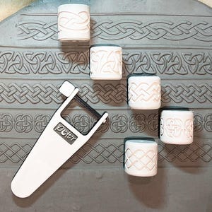 May include: Six white ceramic rollers with Celtic knot designs, used for creating decorative patterns on clay. A white plastic tool with the word "Potter" printed on it is also in the image.