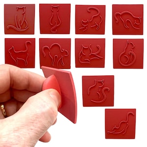 May include: Set of ten red rubber stamps featuring different cat designs. The stamps are square and have a raised design. The stamps are arranged in a grid pattern on a white background.