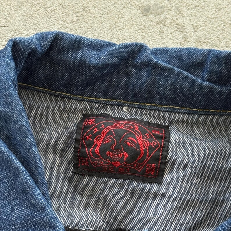 May include: Close-up of a blue denim jacket. A black and red label is sewn onto the jacket. The label features a red illustration of a face and text in a foreign language.