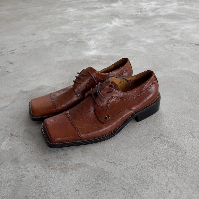 Puede incluir: Un par de zapatos oxford de cuero marr&oacute;n con punteras cuadradas y cierre de cordones. Los zapatos tienen un patr&oacute;n texturizado, en relieve y un brillo sutil.