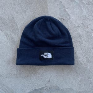 Op de afbeelding: Een marineblauwe beanie met een omgeslagen rand. Een wit en zwart rechthoekig label met "THE NORTH FACE" is gecentreerd op de rand. De hoed is gemaakt van een zacht, gebreid materiaal.