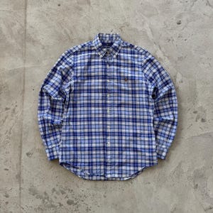 Puede incluir: Camisa de manga larga a cuadros azules y blancos. Presenta un cuello abotonado y puños abotonados. Se distingue un pequeño logo bordado en el pecho. La tela parece ligera y el corte es clásico.