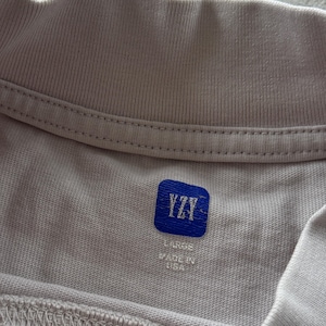 May include: A light beige t-shirt featuring a ribbed crew neckline. The interior neck label is a blue square patch with the white text YZY. Below the patch, the text reads LARGE and MADE IN USA in white, clearly visible against the fabric.