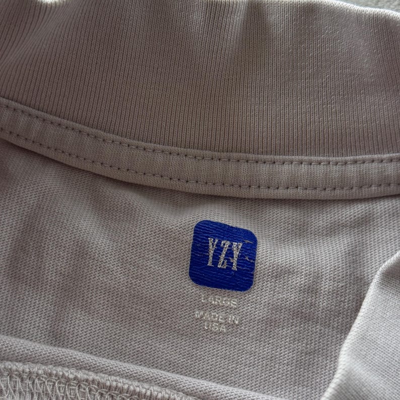 May include: A light beige t-shirt featuring a ribbed crew neckline. The interior neck label is a blue square patch with the white text YZY. Below the patch, the text reads LARGE and MADE IN USA in white, clearly visible against the fabric.