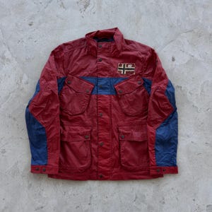 May include: A red and blue jacket with a button-up front, four front pockets, and a small patch with a flag design. The jacket has blue accents on the sleeves and across the chest.