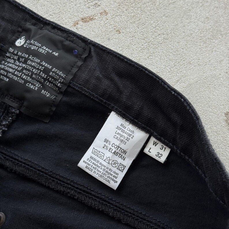 May include: Close-up of black denim jeans with tags. The tags display information about the jeans, including the brand, size, and material composition. The jeans are labeled as size W31 L32. The fabric is 98% cotton and 2% elastane.