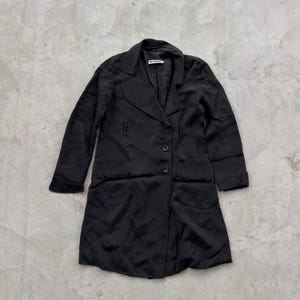 May include: A black long-sleeved coat featuring a notched collar, a two-button front closure, and two large rounded patch pockets at the hips. The interior neck label displays the text JIL SANDER in white lettering on a black background.
