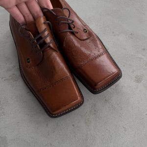 Puede incluir: Un par de zapatos oxford de cuero marr&oacute;n con punteras cuadradas y un patr&oacute;n texturizado similar al de avestruz. Los zapatos tienen cordones y un acento dorado en la punta.