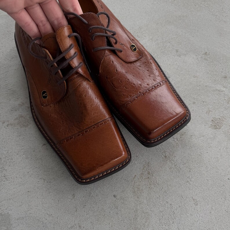Puede incluir: Un par de zapatos oxford de cuero marr&oacute;n con punteras cuadradas y un patr&oacute;n texturizado similar al de avestruz. Los zapatos tienen cordones y un acento dorado en la punta.
