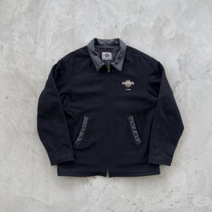 May include: Black jacket with a gray leather collar and Hard Rock Cafe Sydney logo. The jacket has a full zipper, two front pockets, and a small logo patch on the collar. A classic, casual jacket.