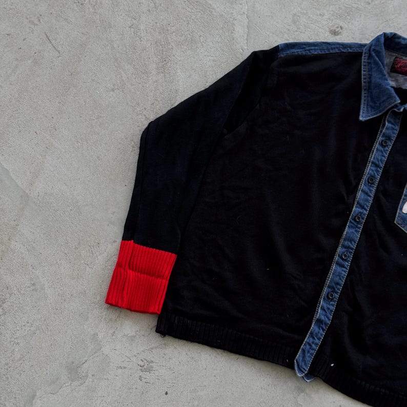 May include: Black shirt with a denim collar and button placket. The long sleeves have red ribbed cuffs. A small white patch is visible on the chest. The shirt is made of a soft, dark fabric.