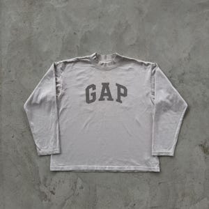 May include: A light gray long-sleeve shirt featuring a mock neck collar and the word GAP printed in a darker gray font across the chest. The garment is constructed with long sleeves and a straight hemline, showcasing a simple and functional design.