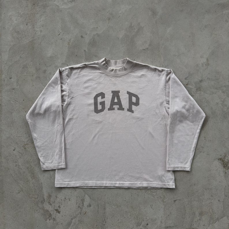May include: A light gray long-sleeve shirt featuring a mock neck collar and the word GAP printed in a darker gray font across the chest. The garment is constructed with long sleeves and a straight hemline, showcasing a simple and functional design.