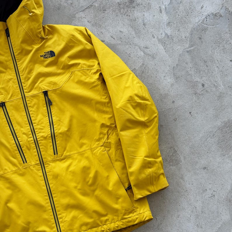 May include: Bright yellow jacket with a black zipper and two black zippered pockets. The North Face logo is visible on the upper left chest. The jacket has a hood and is made of a lightweight material.