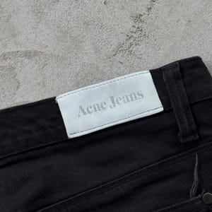May include: Close-up of black denim jeans. The white label on the back of the jeans reads "Acne Jeans" in a gray font. The jeans are made of a dark, solid black material.