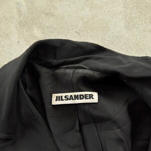 May include: A black jacket with a wide collar and a black interior lining. A rectangular white fabric label is sewn into the inner neckline, featuring the text JILSANDER printed in bold black capital letters across the center of the tag.