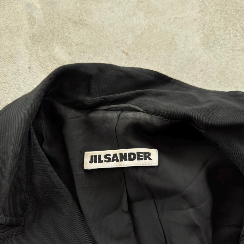 May include: A black jacket with a wide collar and a black interior lining. A rectangular white fabric label is sewn into the inner neckline, featuring the text JILSANDER printed in bold black capital letters across the center of the tag.
