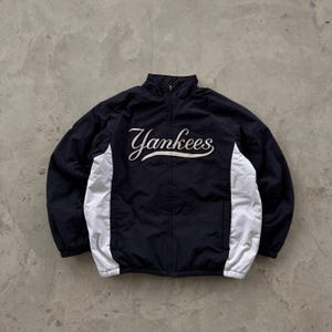 Op de afbeelding: Marineblauwe Yankees-jas met witte zijpanelen en een volledige ritssluiting. Het woord "Yankees" is in witte letters op de borst geborduurd. Het heeft een hoge kraag.