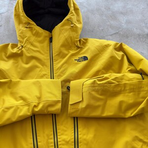 May include: A bright yellow hooded jacket with a black interior and a black zipper. The jacket has the logo "THE NORTH FACE" on the chest and the word "DRYVENT" on the sleeve.