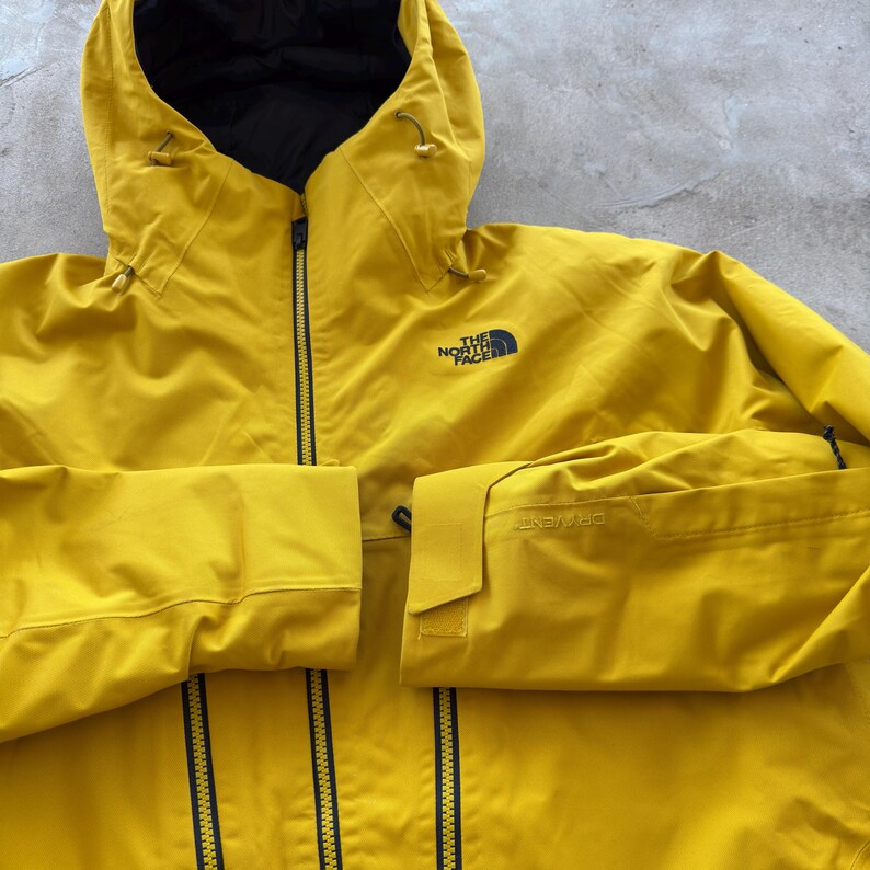 May include: A bright yellow hooded jacket with a black interior and a black zipper. The jacket has the logo "THE NORTH FACE" on the chest and the word "DRYVENT" on the sleeve.