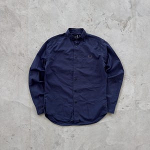 May include: A navy blue button-down shirt with long sleeves. The shirt has a classic collar and a small embroidered emblem on the chest. The shirt is made of a smooth fabric and is buttoned up. The shirt is laid flat on a gray surface.