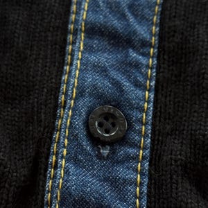 May include: Close-up of a black buttoned shirt with a denim-like blue fabric panel. The panel is stitched with yellow thread. The button is black and has a circular design.