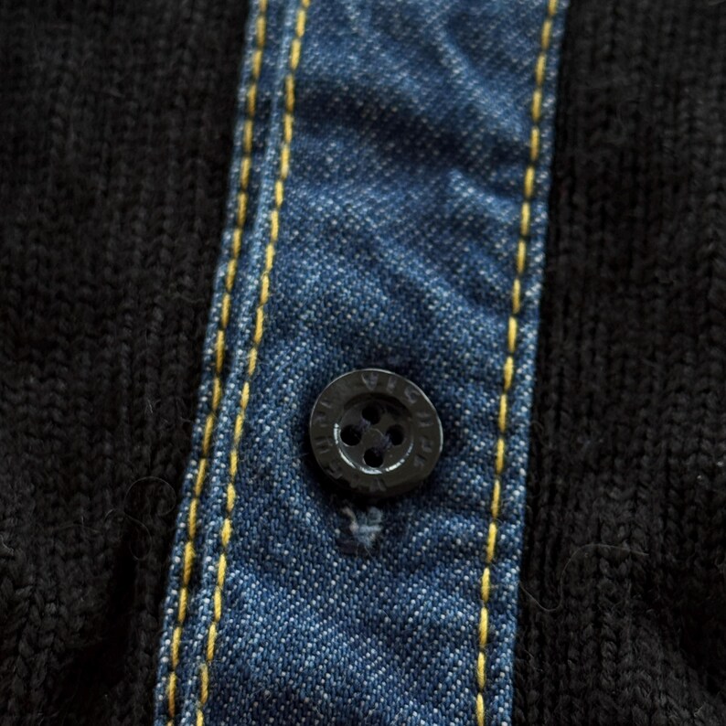 May include: Close-up of a black buttoned shirt with a denim-like blue fabric panel. The panel is stitched with yellow thread. The button is black and has a circular design.
