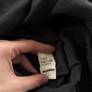 May include: A black garment featuring an interior white fabric care label. The label displays the text 100% RAYON, includes standard laundry care symbols, states MADE IN GERMANY, and features the brand name JIL SANDER in bold black lettering.