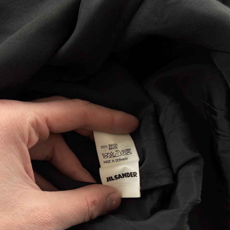 May include: A black garment featuring an interior white fabric care label. The label displays the text 100% RAYON, includes standard laundry care symbols, states MADE IN GERMANY, and features the brand name JIL SANDER in bold black lettering.