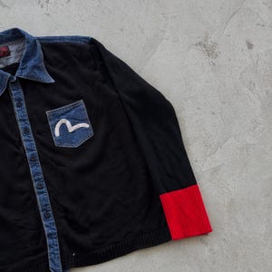 May include: Black button-up shirt with a denim collar and pocket. The pocket has a white wave logo on a denim patch. The sleeve cuff is red. The shirt is a casual style.