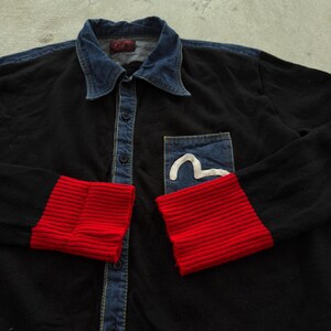 May include: Black button-up shirt with a denim collar and pocket. The cuffs are red ribbed knit. A white curved design is on the denim pocket. The shirt has a vintage look.