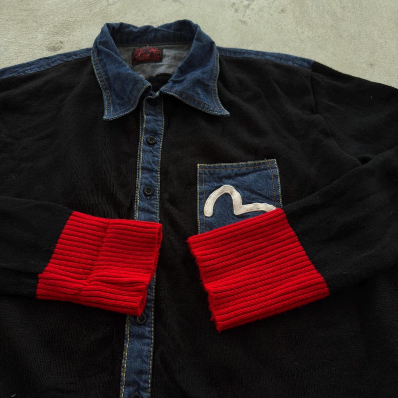 May include: Black button-up shirt with a denim collar and pocket. The cuffs are red ribbed knit. A white curved design is on the denim pocket. The shirt has a vintage look.