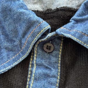 May include: Close-up of a black knit shirt with a denim collar. The collar is a medium blue with yellow stitching. A single dark button fastens the collar.