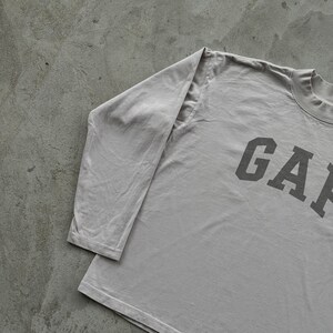 May include: A light gray long-sleeve shirt featuring a mock neck collar and the word GAP printed in large, dark gray block letters across the chest. The garment has long sleeves and a straight hemline, constructed from a solid-colored fabric.