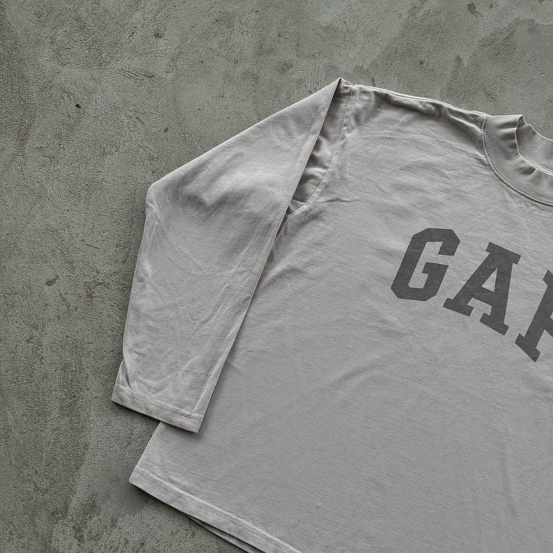 May include: A light gray long-sleeve shirt featuring a mock neck collar and the word GAP printed in large, dark gray block letters across the chest. The garment has long sleeves and a straight hemline, constructed from a solid-colored fabric.