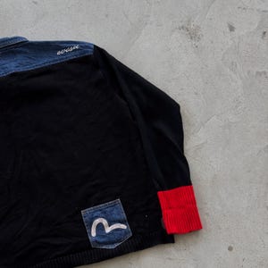 May include: Black long-sleeve shirt with a denim blue shoulder yoke and a red ribbed cuff. A small denim pocket with a white graphic is sewn on the front. The word "evisu" is embroidered in white on the shoulder.