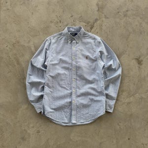Puede incluir: Camisa abotonada a rayas azules y blancas con mangas largas. La camisa tiene cuello abotonado y un pequeño logo marrón en el pecho. La camisa está hecha de tela ligera.