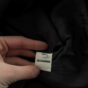 May include: A black garment featuring an interior white fabric tag. The tag displays the text JIL SANDER in black capital letters, with the number 36 printed in a smaller font size directly above the brand name on a separate small white tab.