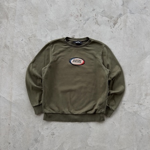 May include: An olive green sweatshirt with a crew neck and long sleeves. It has a Diesel logo patch in a red, white, and blue oval on the chest. The fabric looks soft and slightly worn.