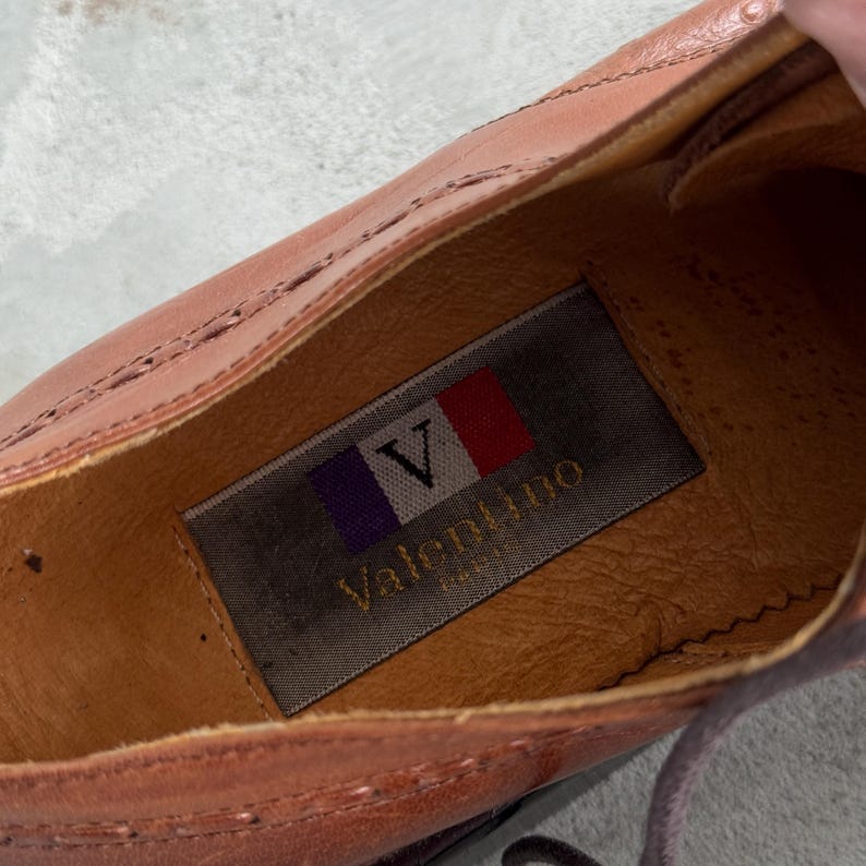 Puede incluir: Primer plano del interior de un zapato de cuero marr&oacute;n con una etiqueta que dice "Valentino" en letras blancas con un dise&ntilde;o de bandera roja, blanca y azul.