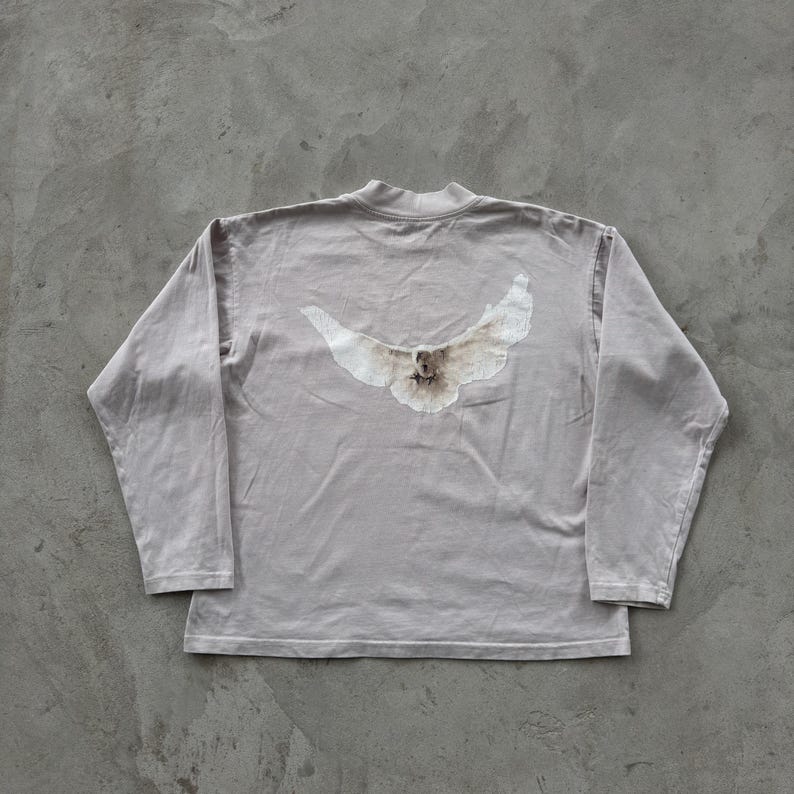 May include: A long-sleeve mock neck shirt in a muted light gray color. The center of the chest features a graphic print of a white bird with its wings spread wide, displaying detailed feathers and a soft, faded appearance against the fabric.