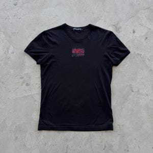 Könnte beinhalten: Schwarzes T-Shirt mit Rundhalsausschnitt und einem kleinen, rechteckigen rot-schwarzen Grafikmuster auf der Brust. Das Shirt hat kurze Ärmel und eine klassische Passform. Der Markenname ist am Ausschnitt sichtbar.