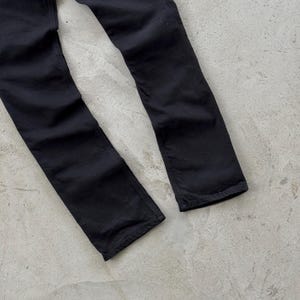 May include: A pair of black denim jeans. The jeans are a solid black color and appear to be straight-legged. The fabric has a slightly worn appearance. The jeans are lying on a light gray surface.