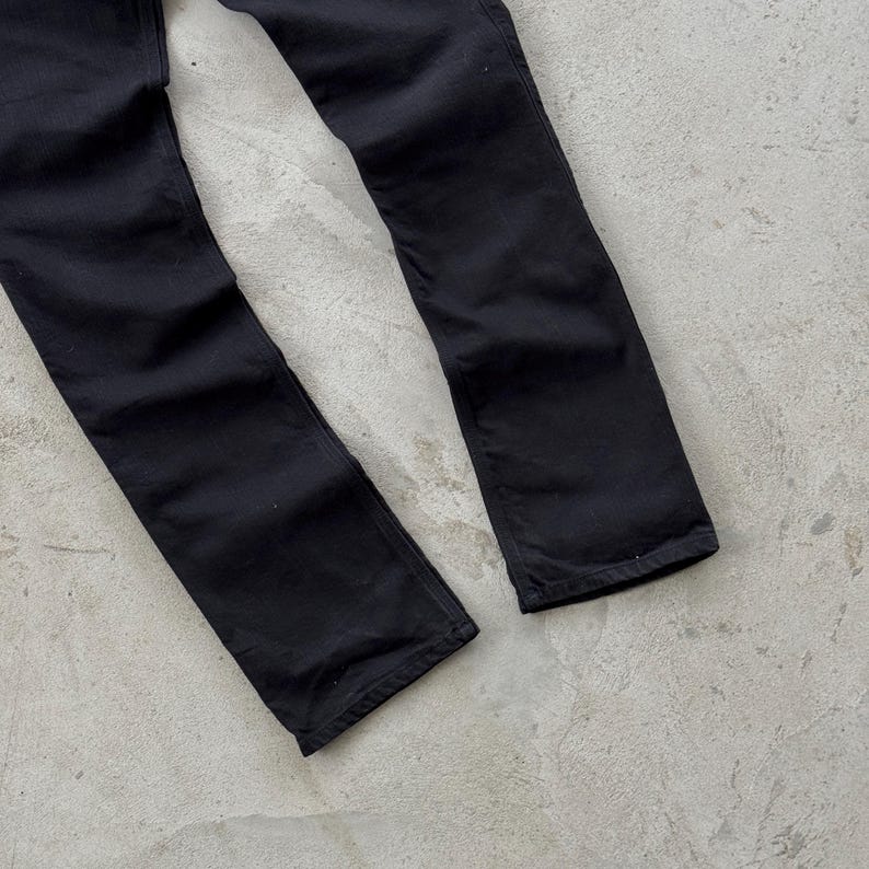May include: A pair of black denim jeans. The jeans are a solid black color and appear to be straight-legged. The fabric has a slightly worn appearance. The jeans are lying on a light gray surface.