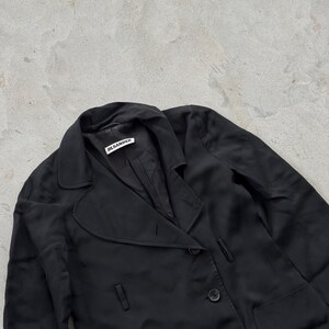 May include: A black blazer with a notched lapel, long sleeves, and a button-front closure. The interior collar features a white fabric label with the black text JIL SANDER. The garment is constructed from a smooth, lightweight, dark fabric.