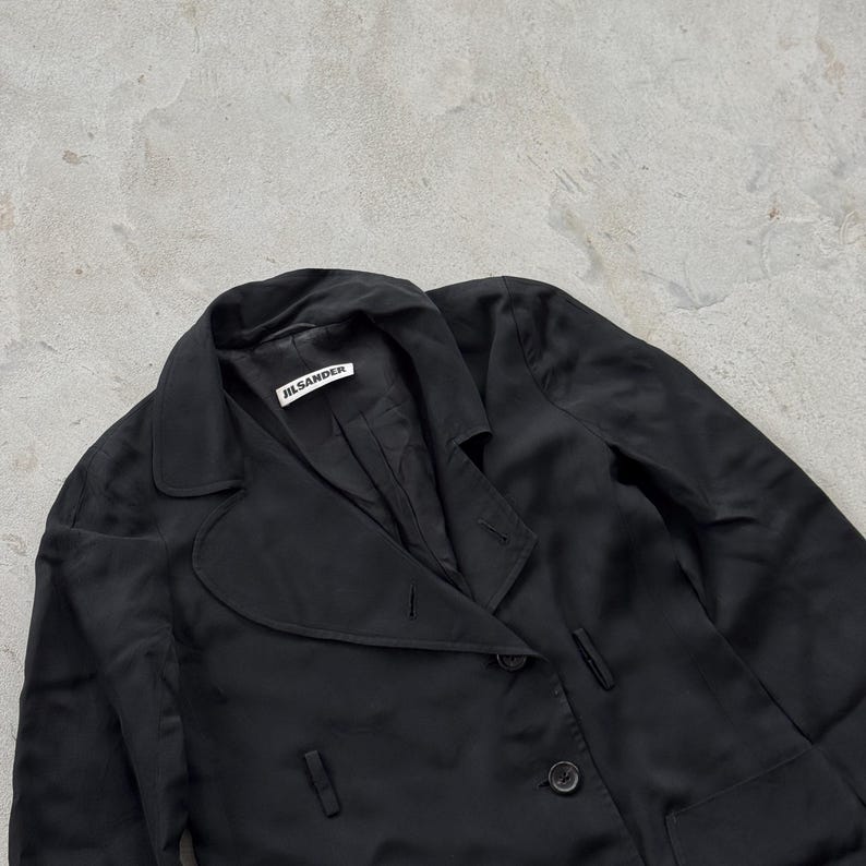 May include: A black blazer with a notched lapel, long sleeves, and a button-front closure. The interior collar features a white fabric label with the black text JIL SANDER. The garment is constructed from a smooth, lightweight, dark fabric.