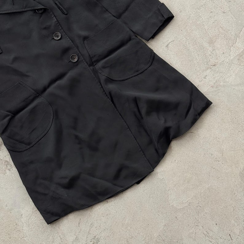 May include: A black coat featuring a collared neckline, a front button closure with three visible dark buttons, long sleeves with cuffs, and two large rounded patch pockets positioned on the lower front section of the garment.